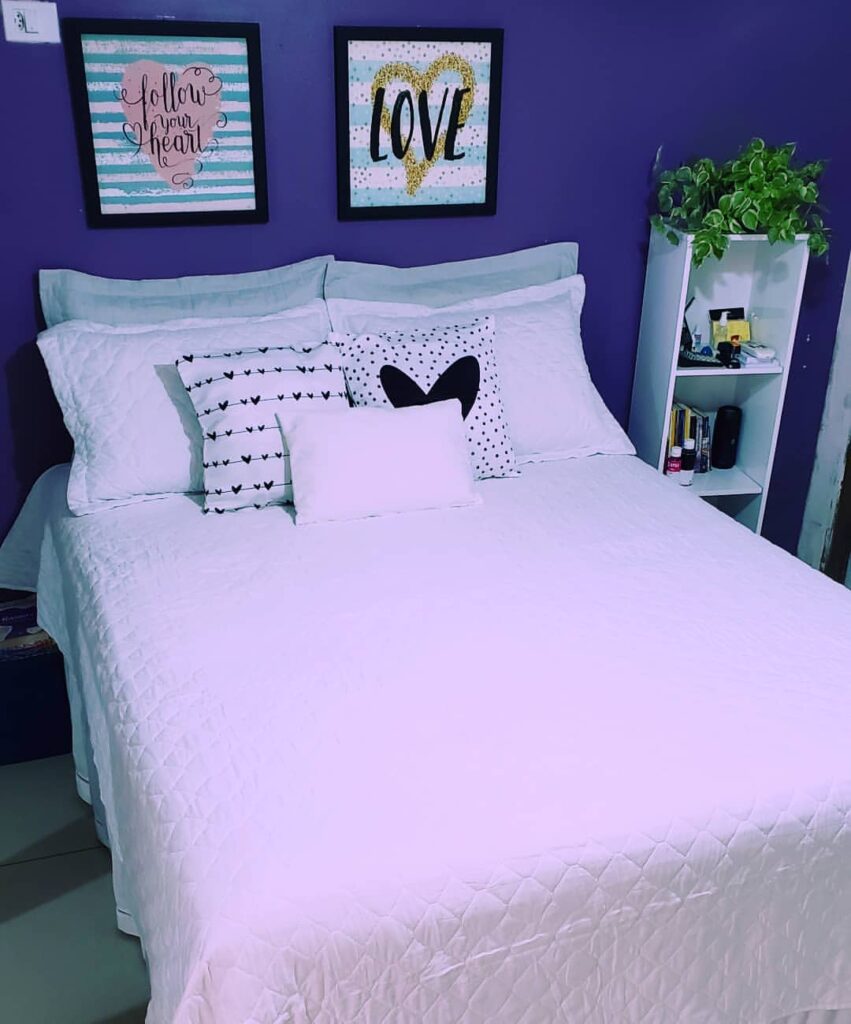 Quarto com parede azul e quadros, cama posta branca com detalhes de coração nas almofadas