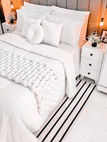 Quarto de casal com cama postra branca com várias almofadas, colcha e manta