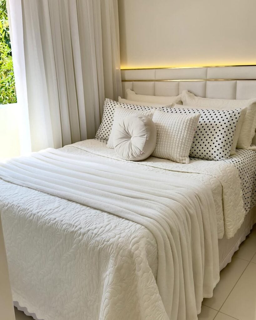 Cama posta branca com almofadas, cobreleito, manta e detalhe dourado na cabeceira