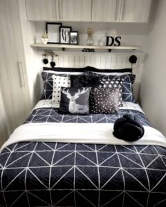 Quarto com cabeceira branca em lambri, prateleira acima com quadrinhos e cama posta com várias almofadas escuras