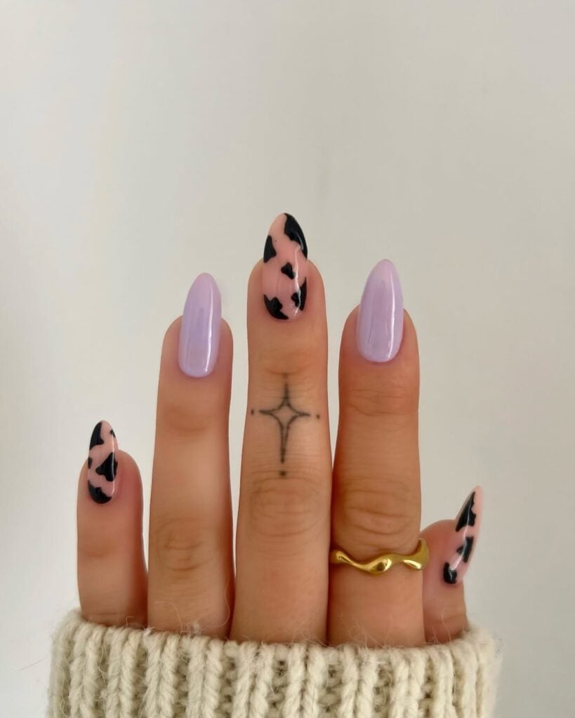 Mão de pele clara, com anel dourado e unha amendoada lilás com cow print