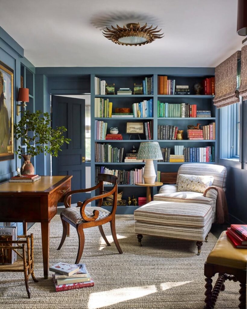 Ambiente com estante azul com livros, poltrona com peseira, escrivaninha de madeira com cadeira ornamentada