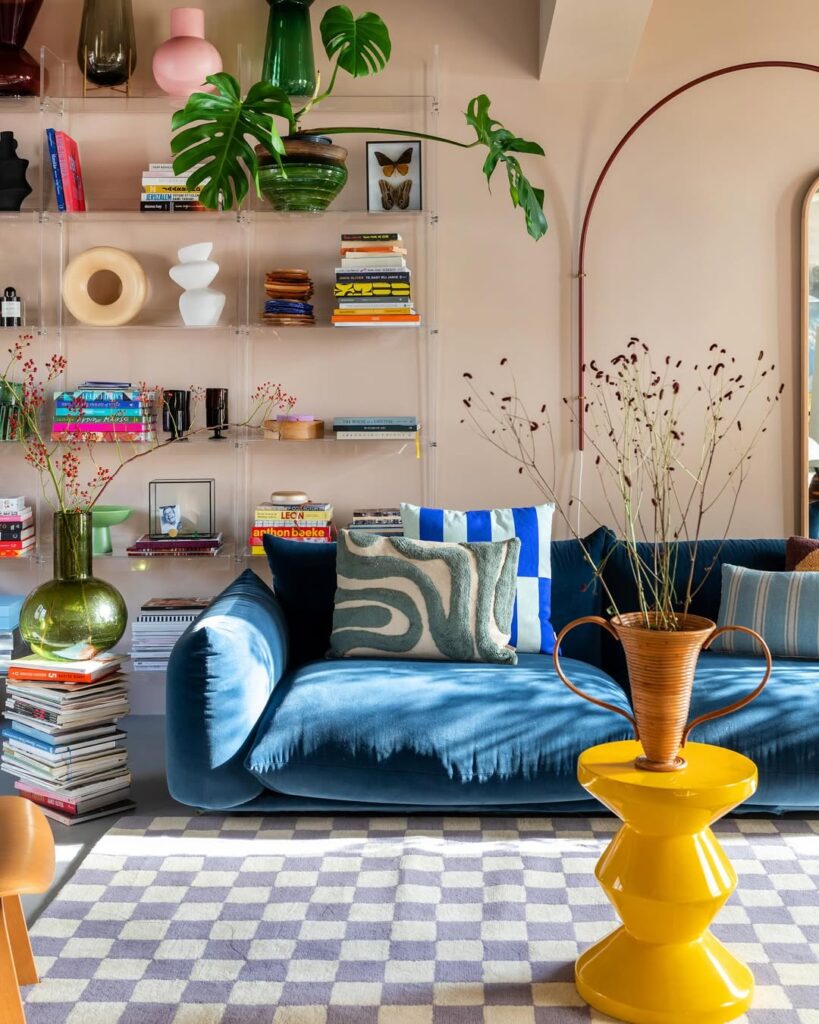 Sala de estar com sofá azul em veludo, tapete quadriculado, estante com objetos e livros e mesa de centro amarela