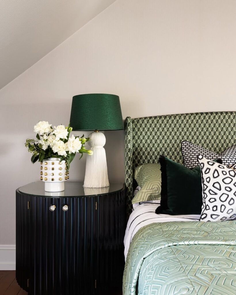 Quarto com mesa de cabeceira escura com vaso de flores brancas e abajur com base branca e cúpula verde