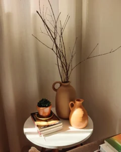 Mesa de canto branca com livros, jarro de barro com galhos secos