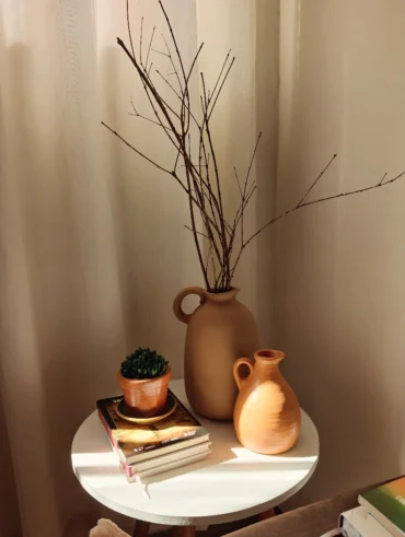 Mesa de canto branca com livros, jarro de barro com galhos secos