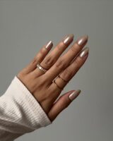 Mão de pele clara, com anéis em dourado e unha amendoada neutra e perolada