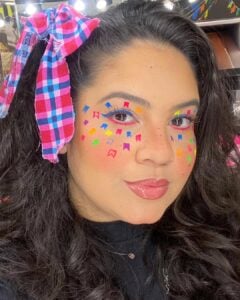 Mulher de pele clara, com cabelo escuro, laço no cabelo e maquiagem colorida com bandeirinhas