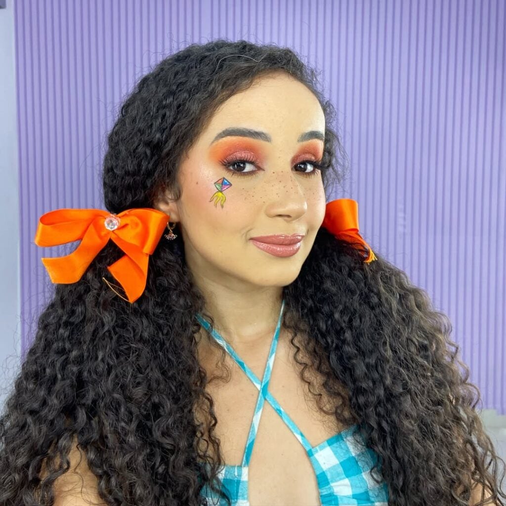 Mulher de pele 'média', com cabelo crespo preso com laços e maquiagem de festa junina
