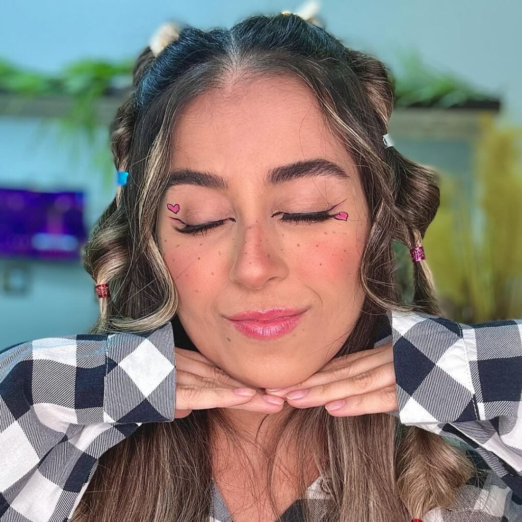 Mulher de pele clara, com camisa xadrez, cabelo loiro preso e maquiagem junina com corações