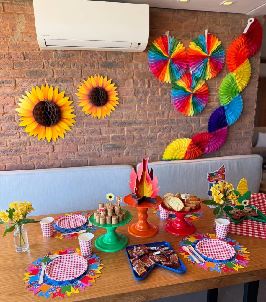 Mesa de madeira com decoração junina, pratinhos xadrez, fogueira de papel e enfeites coloridos na parede