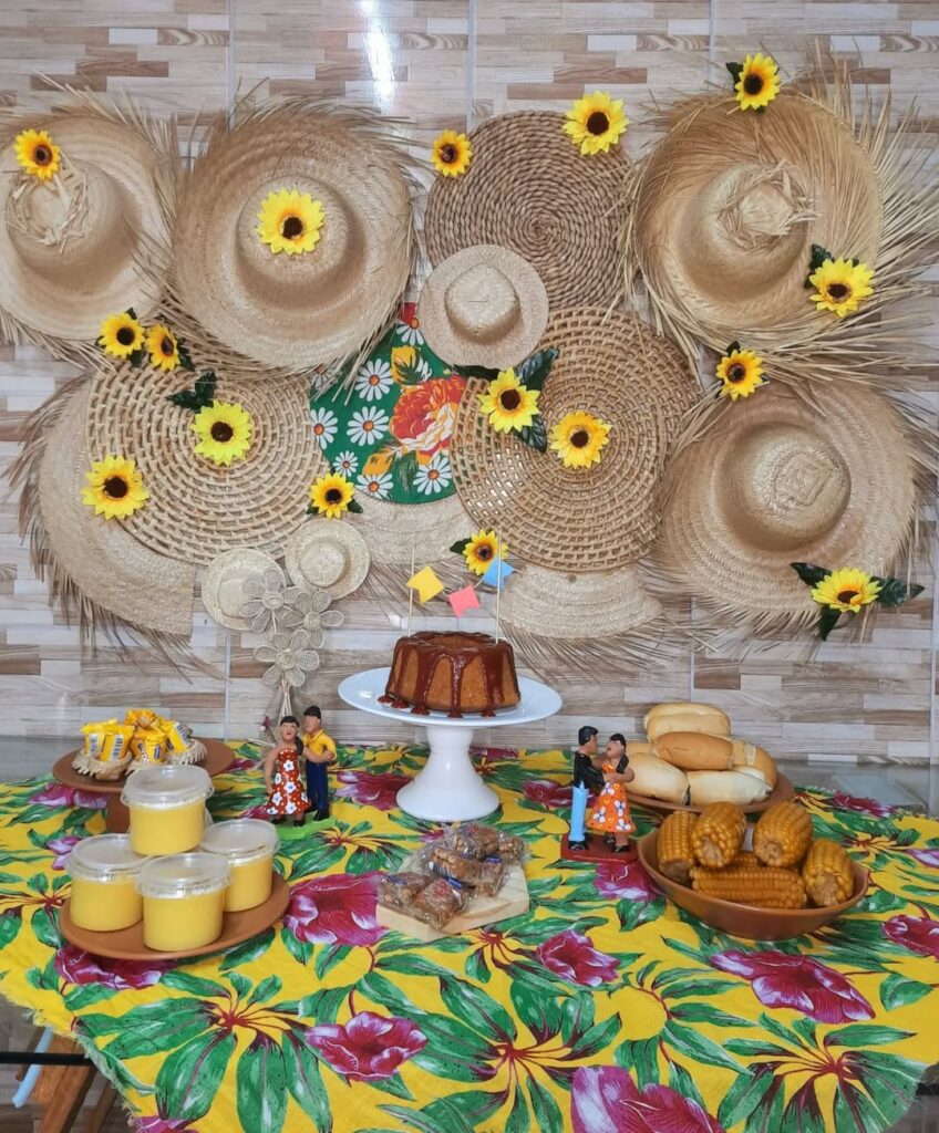 Mesa junina com tecido de chita, bolo com bandeirinhas e chapéus de palha na parede