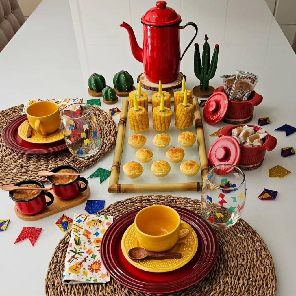 Mesa posta com sousplat de palha, pratos vermelhos, xícaras e pires amarelos, copos com bandeirinhas e bule vermelho