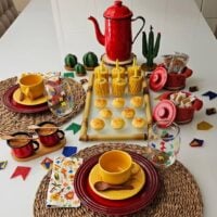 Mesa posta com sousplat de palha, pratos vermelhos, xícaras e pires amarelos, copos com bandeirinhas e bule vermelho