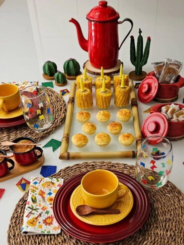 Mesa posta com sousplat de palha, pratos vermelhos, xícaras e pires amarelos, copos com bandeirinhas e bule vermelho