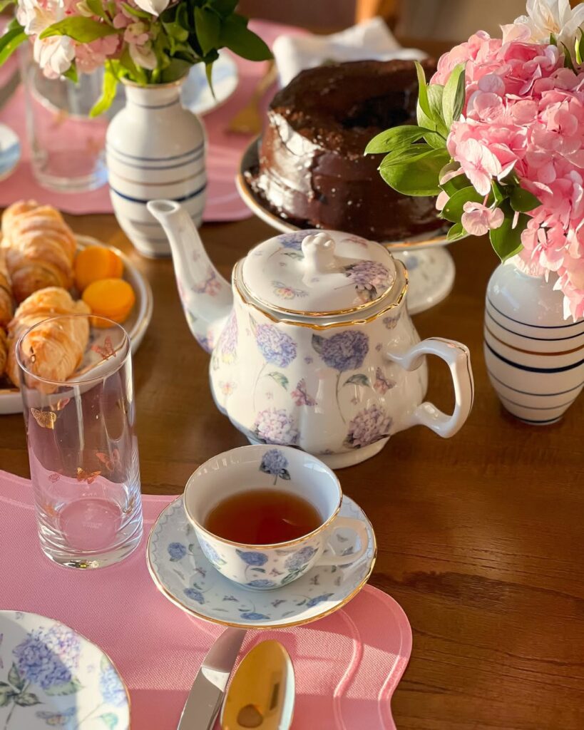 Bule de porcelana com flores e borda dourada, bolo de chocolate e vaso com flores rosas