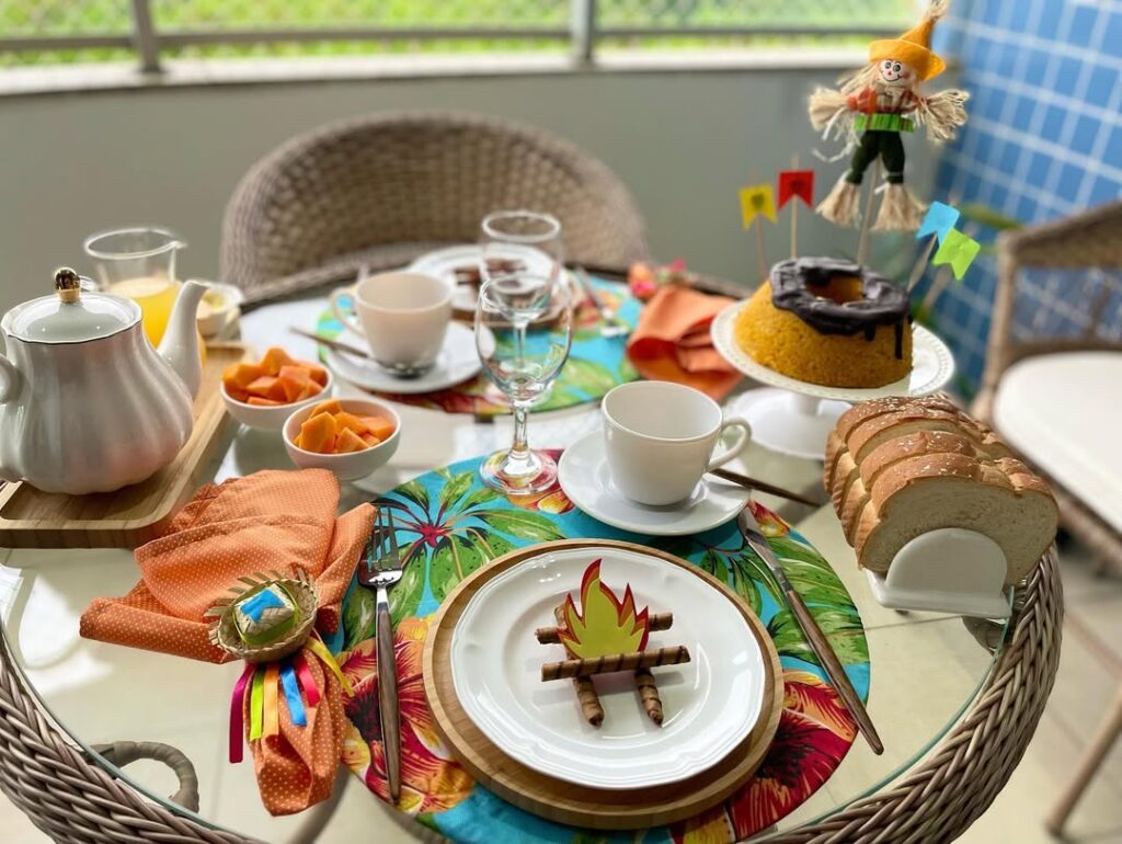 Mesa com tampo de vidro decorada com sousplat colorido, guardanapo com anel de mini chapéu de palha, bolo de milho com bandeirinhas