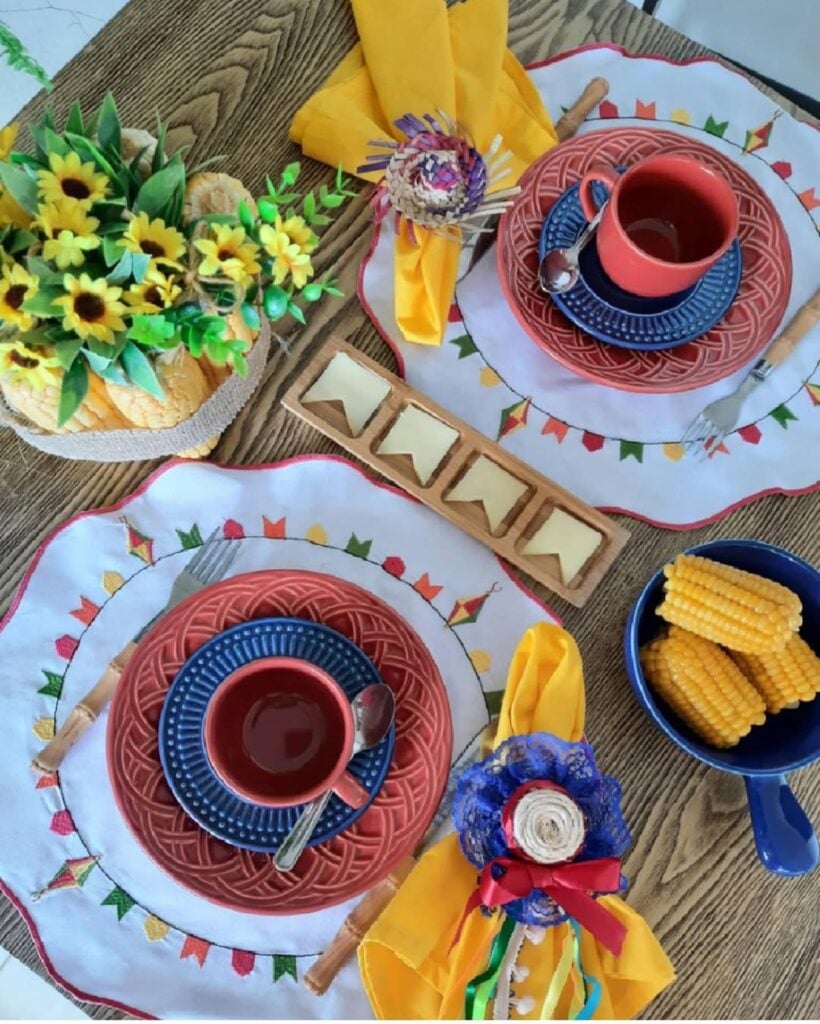 Mesa posta junina com sousplat branco, borda rosa, bandeirinhas e balões bordados