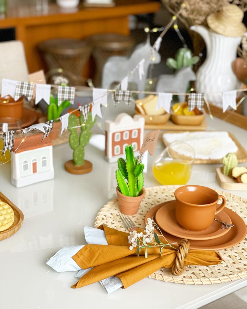 Mesa posta junina com louça caramelo, casinhas, cactus e bandeirinhas