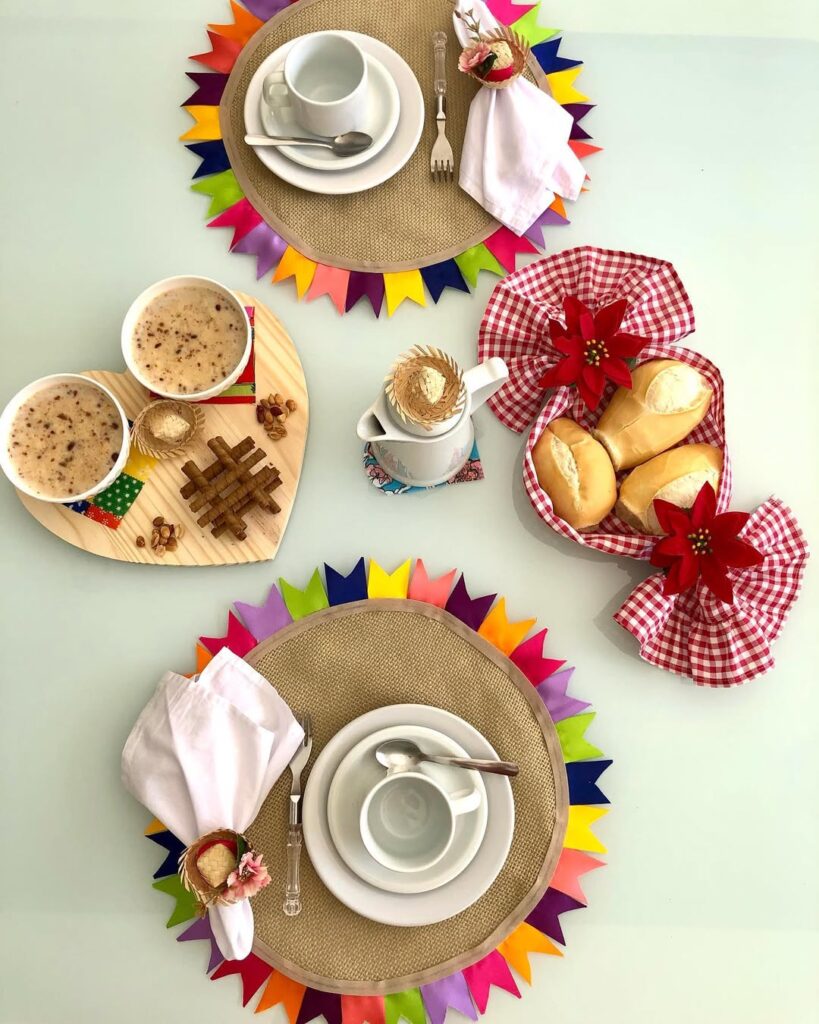 Mesa posta junina com sousplata de juta e bandeirinhas coloridas ao redor, cestinho de pães e tábua em formado de coração