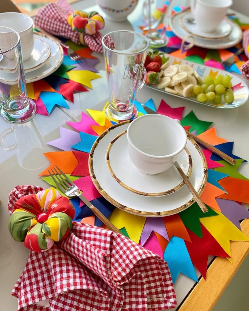Mesa posta com louça branca, sousplat colorido com bandeirinhas e guardanapos xadrez