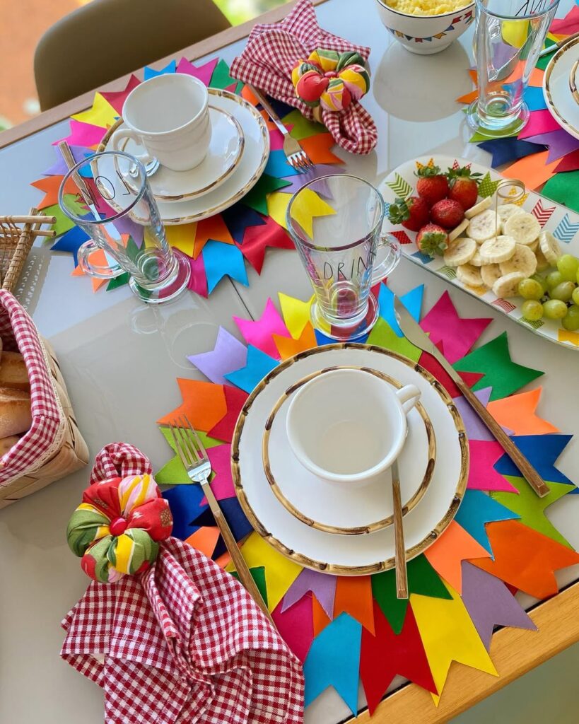 Mesa posta junina com sousplat com bandeirinhas coloridas, guardanapos xadrez e frutas