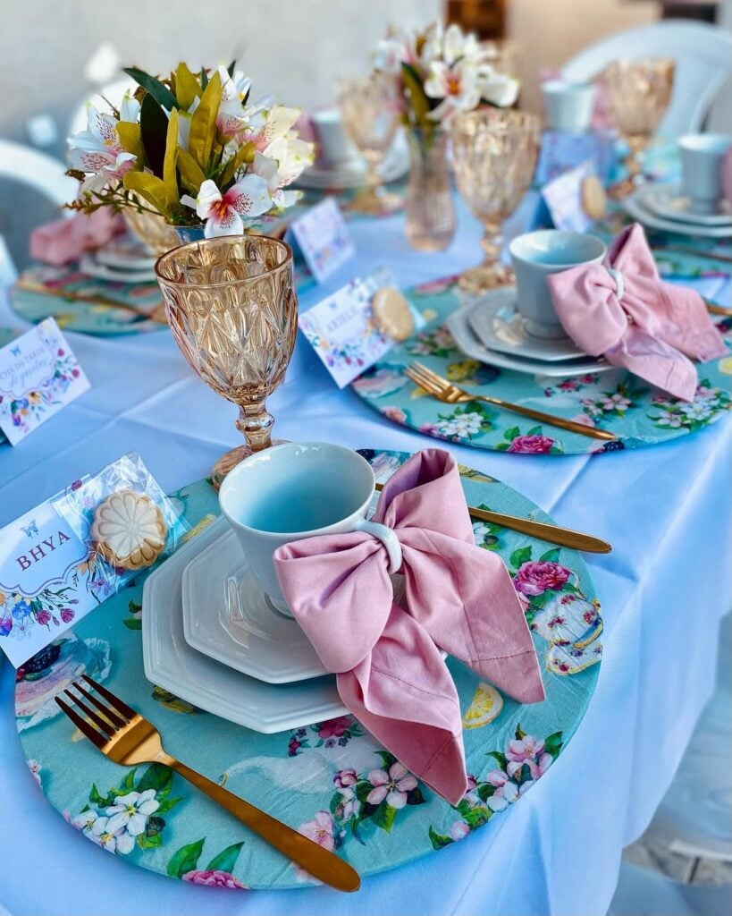 Mesa posta de chá com toalha azul, sousplats floridos, xícaras brancas com laços rosa