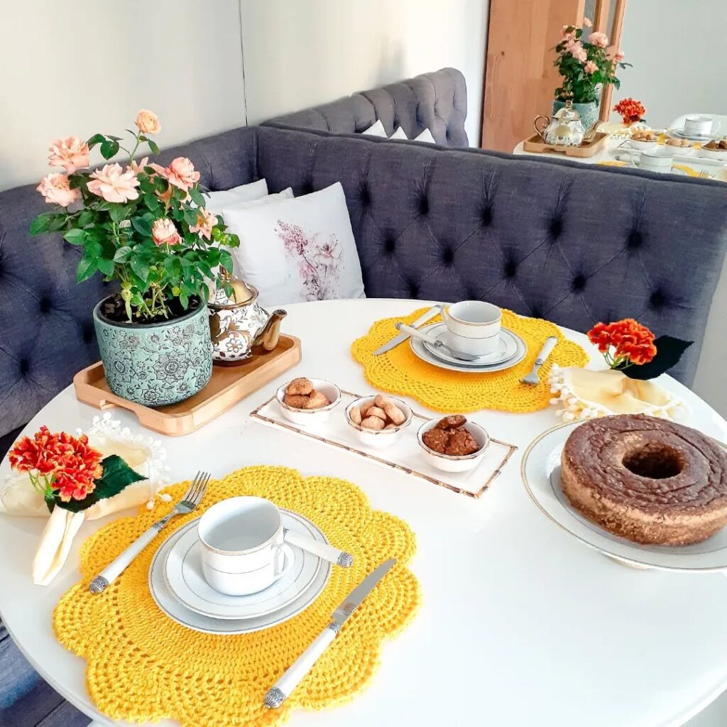 Mesa branca redonda com sousplats amarelos, louças brancas, bolo e flores