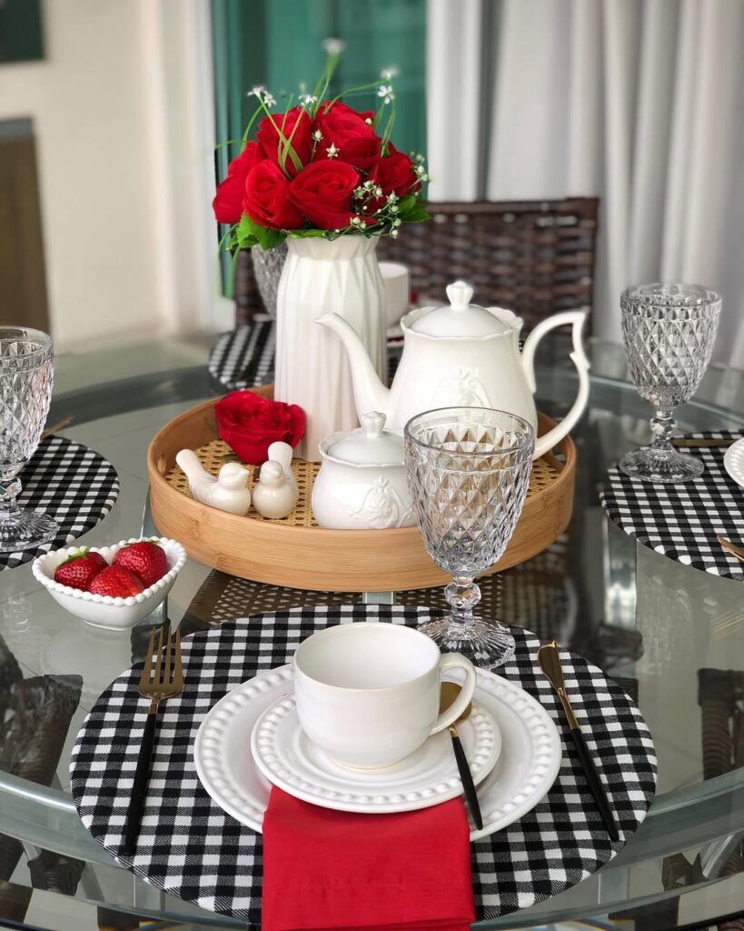 Mesa com sousplats xadrez em preto e branco, louças brancas, bandeja de bambu com bule e rosas vermelhas