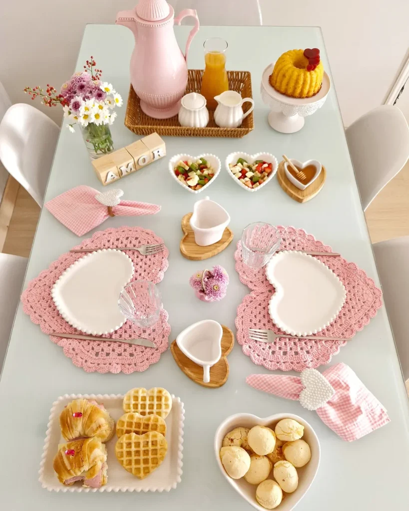 Mesa posta com jogo americano de crochê rosa, pratinhos em formato de coração, vasinho com flores, bolo e pães de queijo