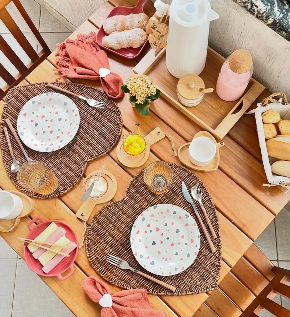Mesa de madeira quadrada com sousplat em formato de coração com pratos brancos e corações rosa e cinza
