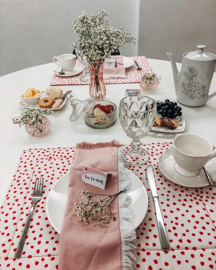 Mesa branca posta com jogo americano em branco e vermelho, guardanapo rosa e vaso com florzinhas brancas
