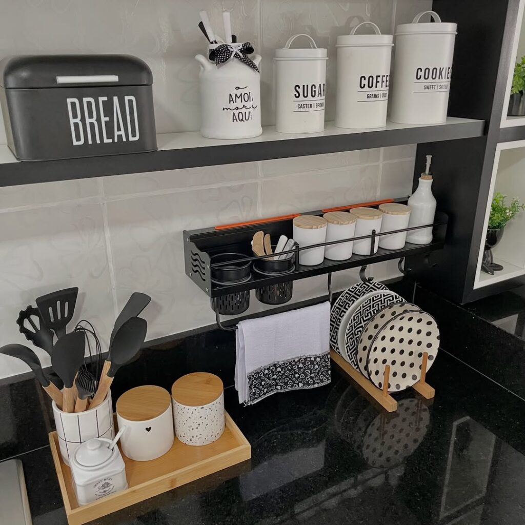 Cozinha com revestimento branco, bancada preta, prateleira com potes para mantimentos e suporte para temperos