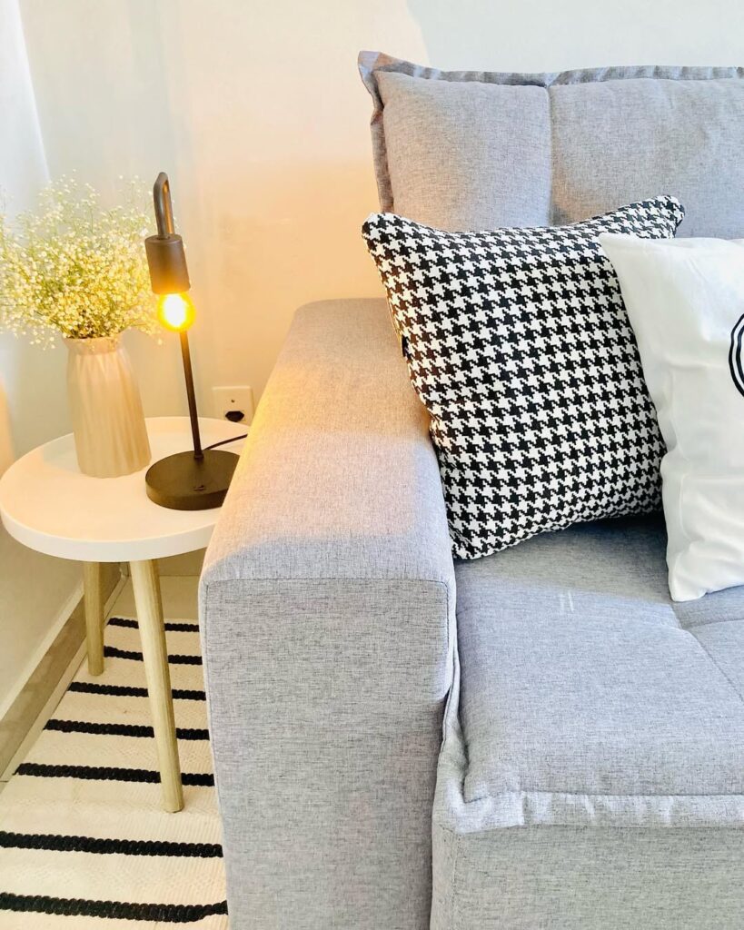 Sala de estar com sofá cinza, mesinha lateral branca com vasinho de flores e luminária
