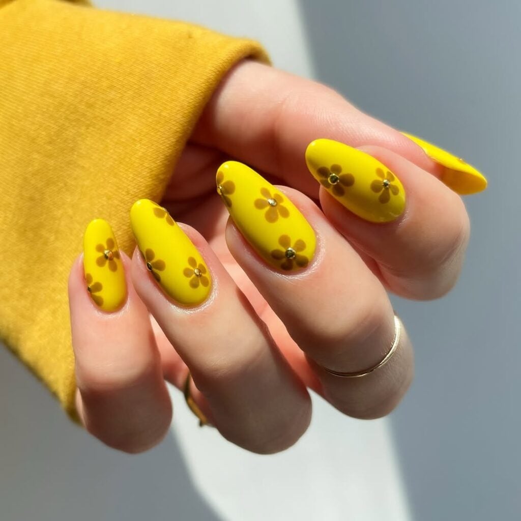 Mão de pele clara, com anéis em dourado e unha amendoada amarela com flores