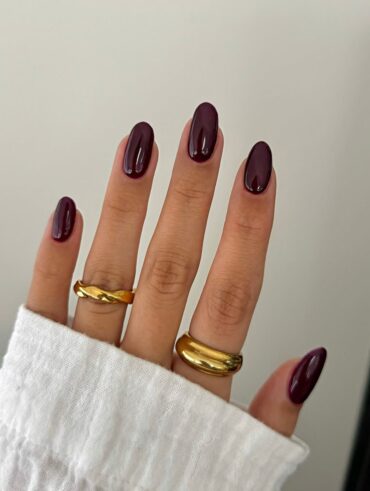 Mão de pele clara, com anéis em dourado e unha amendoada burgundy