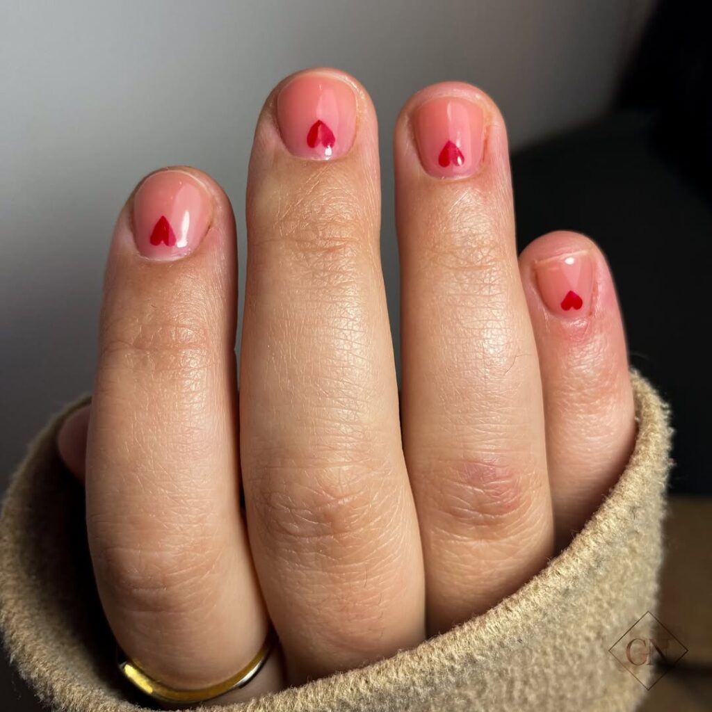 Mão de pele clara, com anel dourado e unha curta rosada com coração vermelho