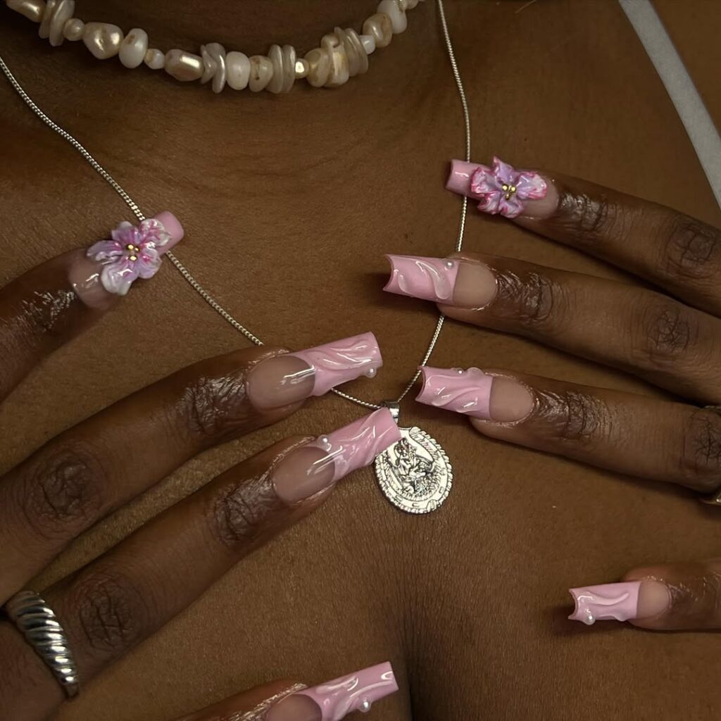 Mãos de pele preta, com acessórios em prata e unha quadrada com inglesinha rosa-clara, decoração em alto relevo e com flor
