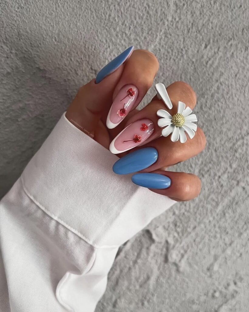 Mão de pele clara, com anel de margarida e unha amendoada azul-claro com francesinha e flores encapsuladas