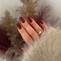 Mão de pele clara, com anel dourado e unha quadrada burgundy