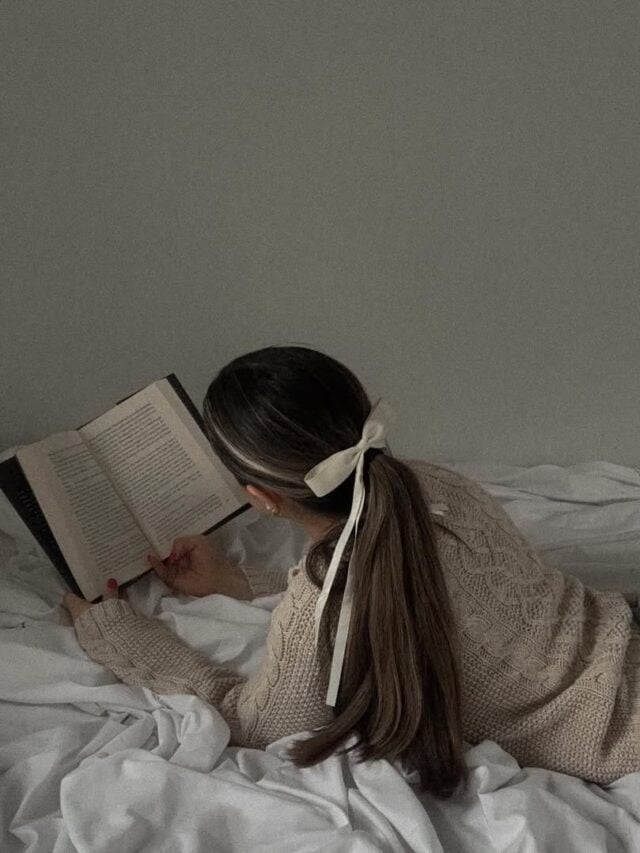 Mulher de pele clara, com cabelo preso. Ela está lendo na cama