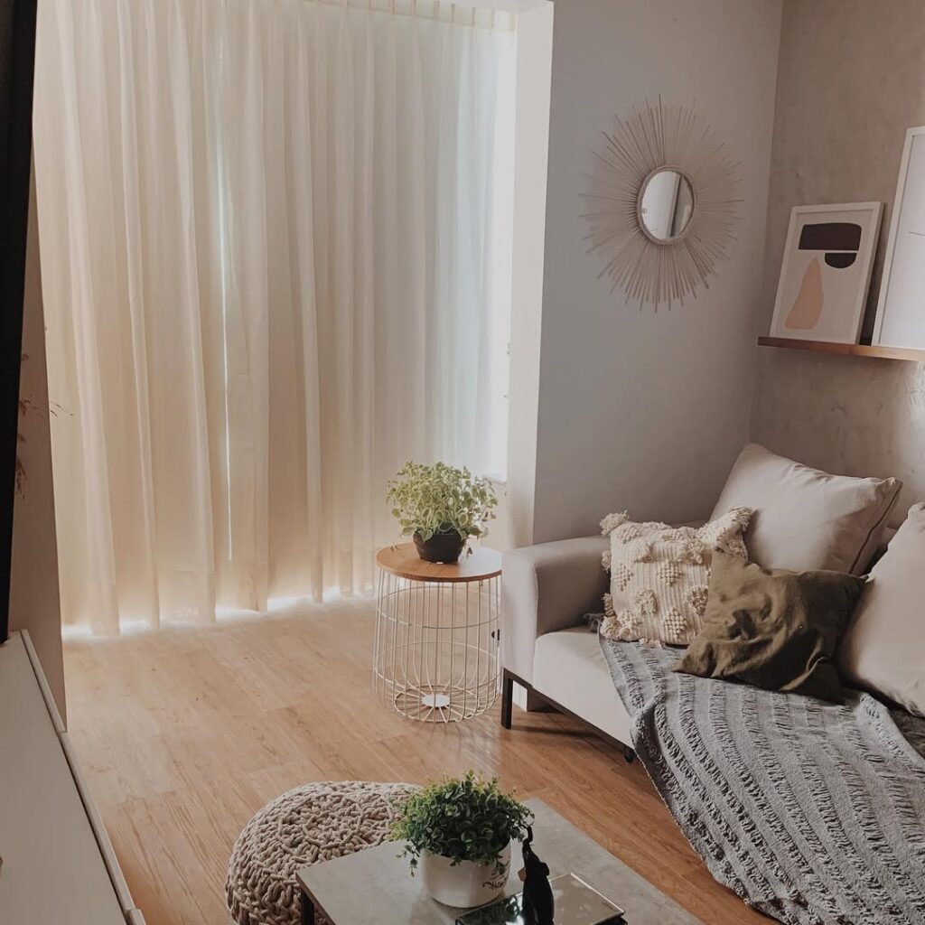 Sala de estar com paredes e sofá em cor clara, espelho de sol e prateleira com quadros