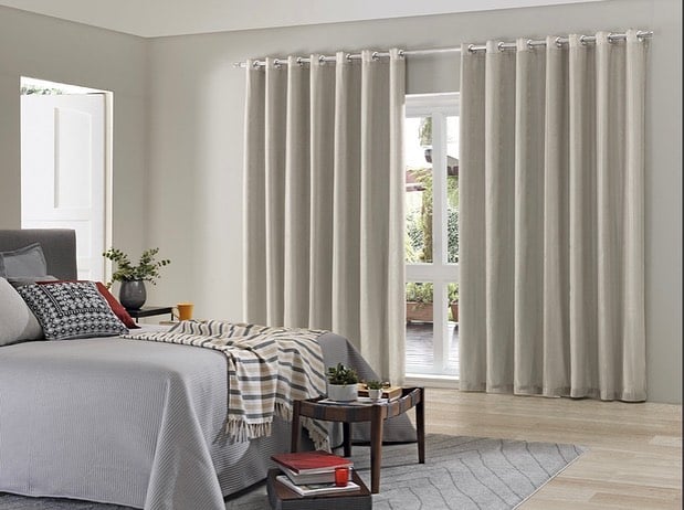 Quarto com mesinha de madeira aos pés da cama, tapete cinza e cortina clara