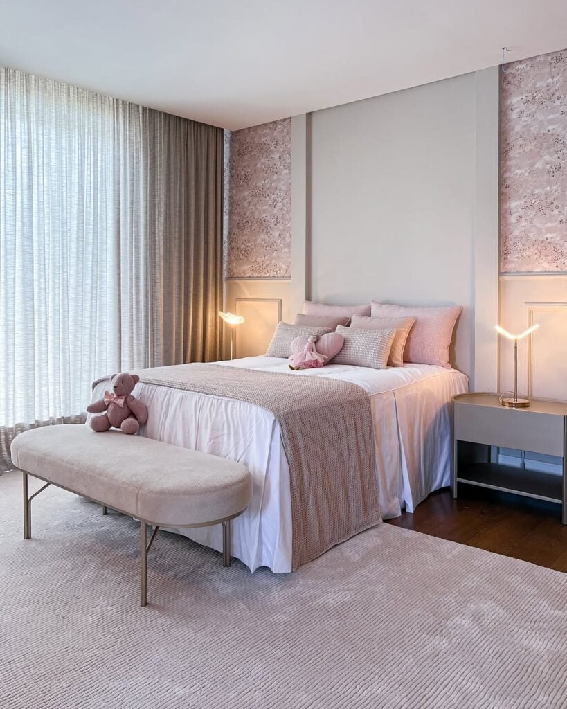 Quarto em tons rosados e branco com almofadas, manta e banco aos pés da cama