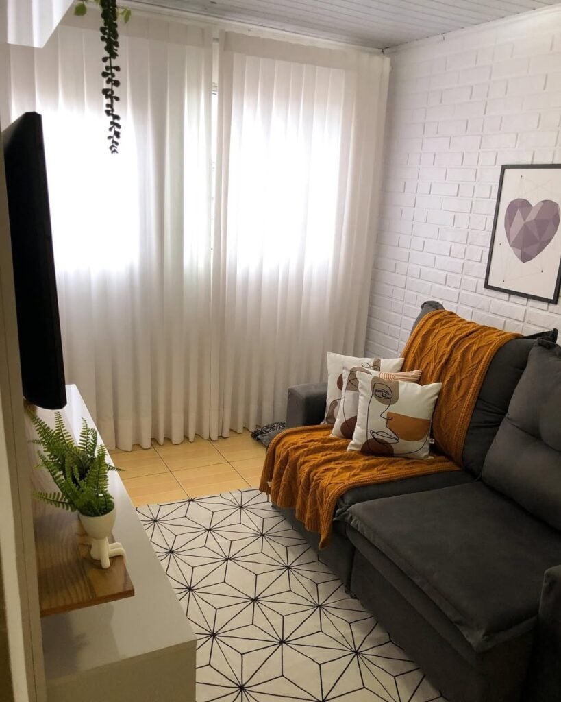 Sala de estar com parede de tijolinhos brancos, sofá cinza em veludo e cortina branca