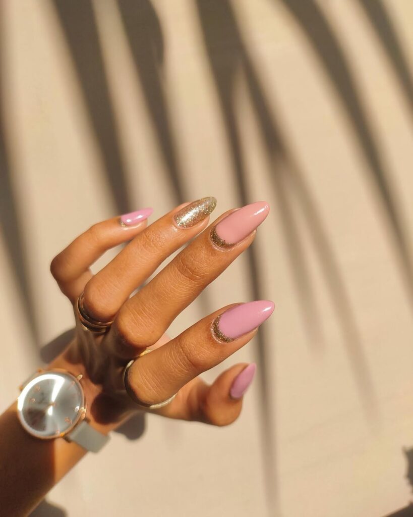 Mão de pele clara, com anéis em dourado e unha amendoada rosa com francesinha invertida brilhosa