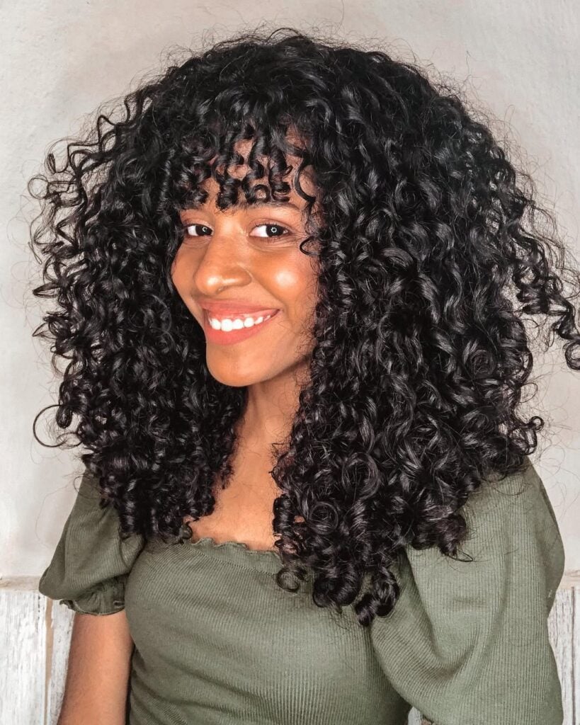Mulher de pele preta, com blusa verde e cabelo preto cacheado com franja reta