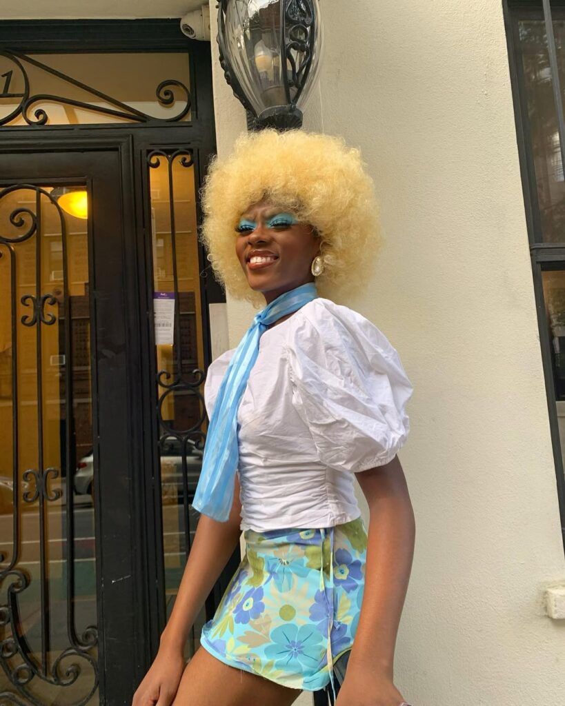 Mulher de pele preta, com blusa branca, saia estampada, lenço, cabelo crespo loiro e maquiagem com sombra azul 