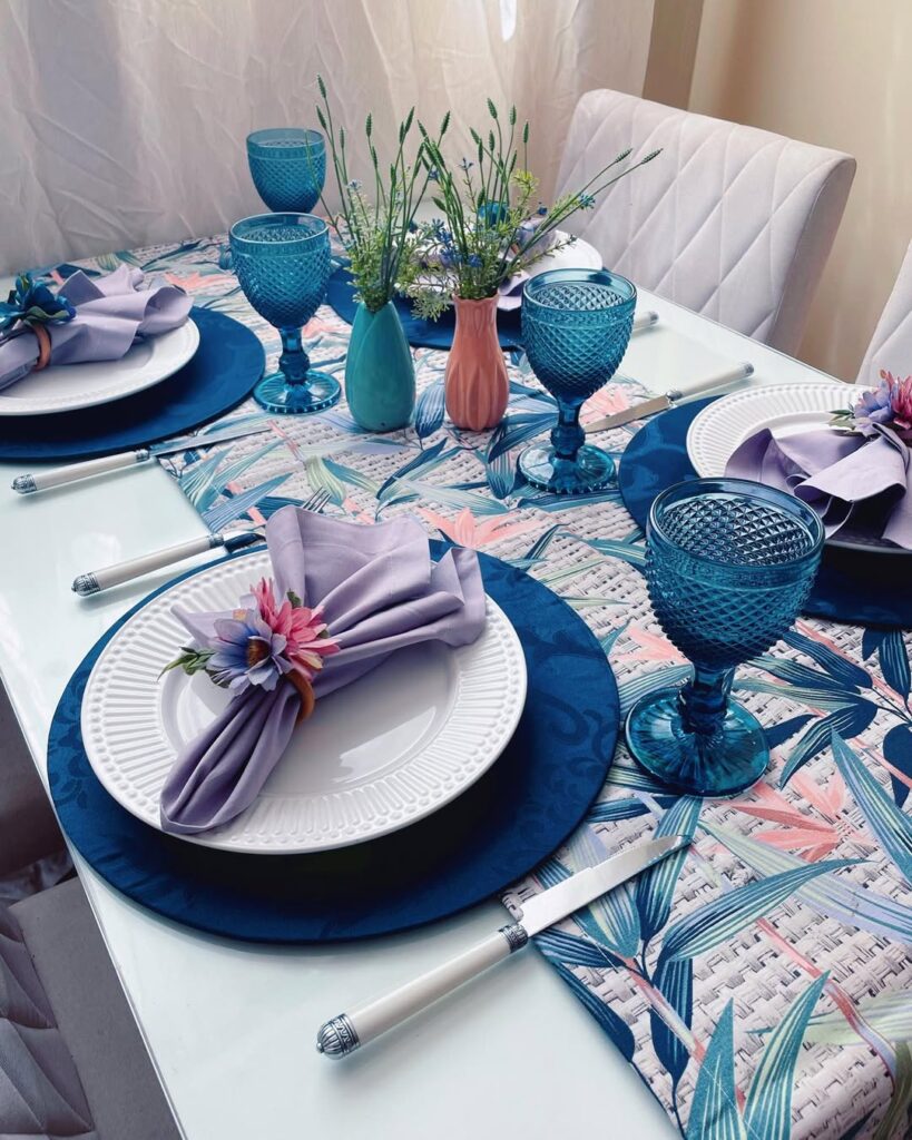 Mesa posta com trilho floral, sousplats azuis, pratos brancos e taças azuis