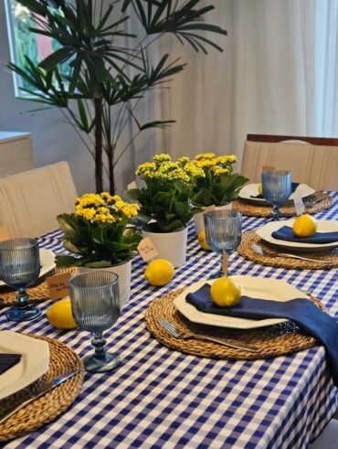 Mesa posta com toalha xadrez em branco e azul, sousplats de palha, pratos brancos, guardanapos azuis e flores amarelas